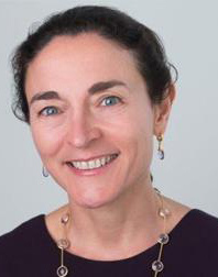 A person with dark hair smiles at the camera, wearing a purple top and a necklace against a plain background.